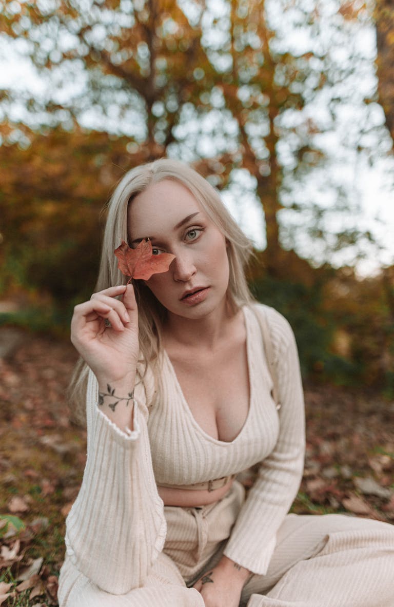 Blonde woman in a sweater holding a leaf in a vibrant autumn forest scene.
