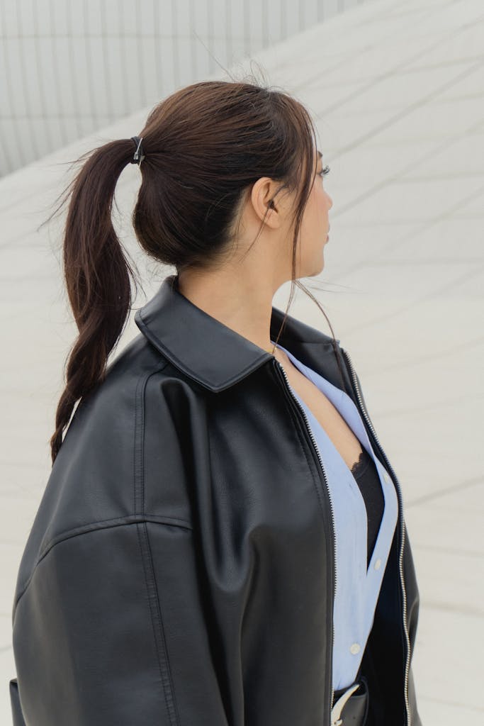 Stylish woman wearing a black leather jacket looks away outside an architectural building.