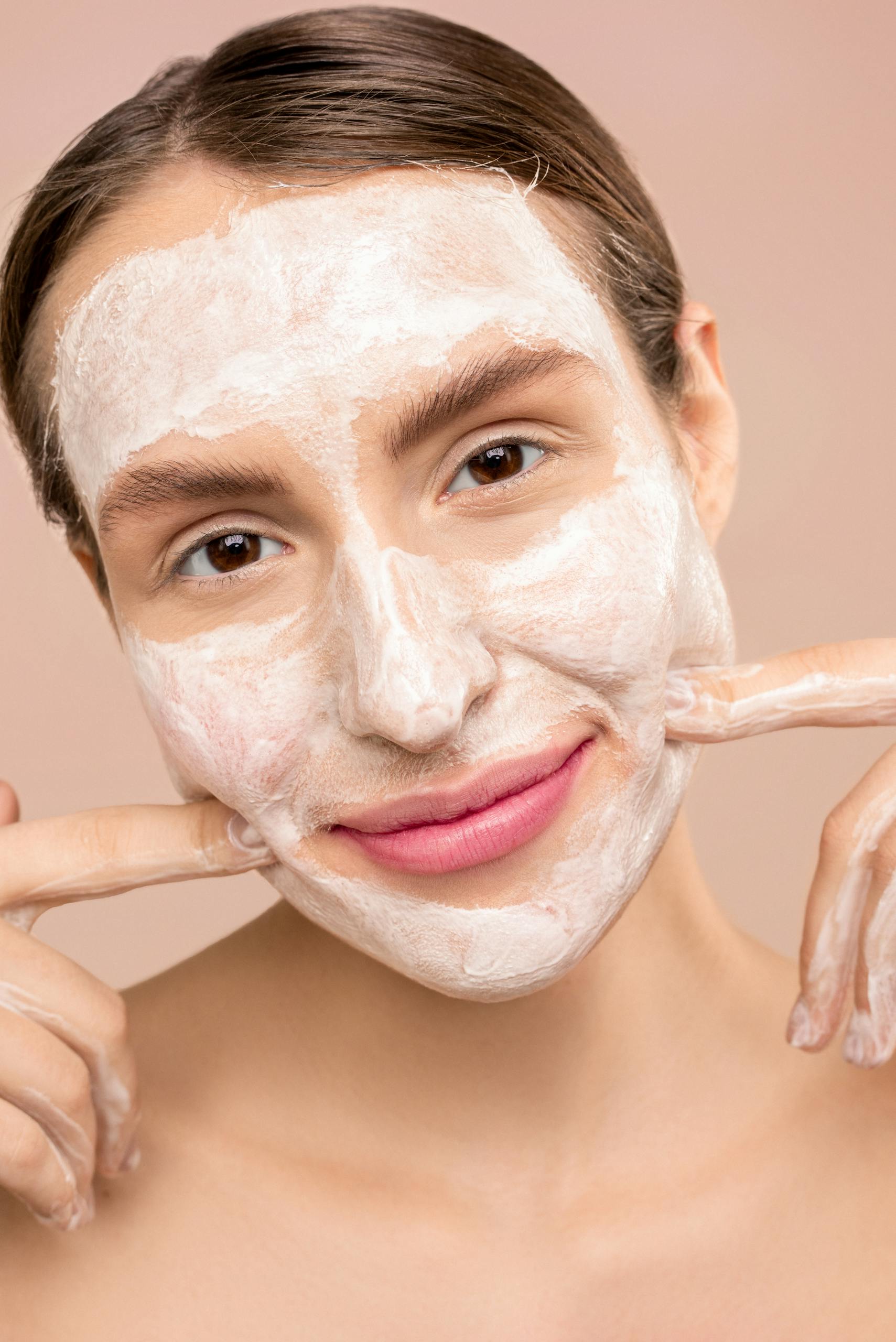 Woman with white facial foam smiling, showcasing skincare routine indoors.